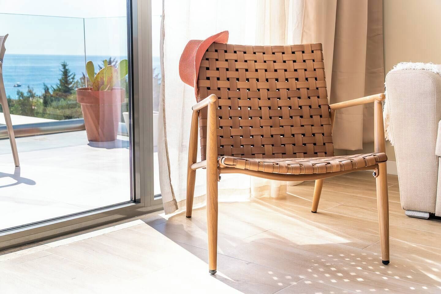 A wicker chair with a cushion, positioned near a sliding glass door with a view of the ocean and potted plants.
