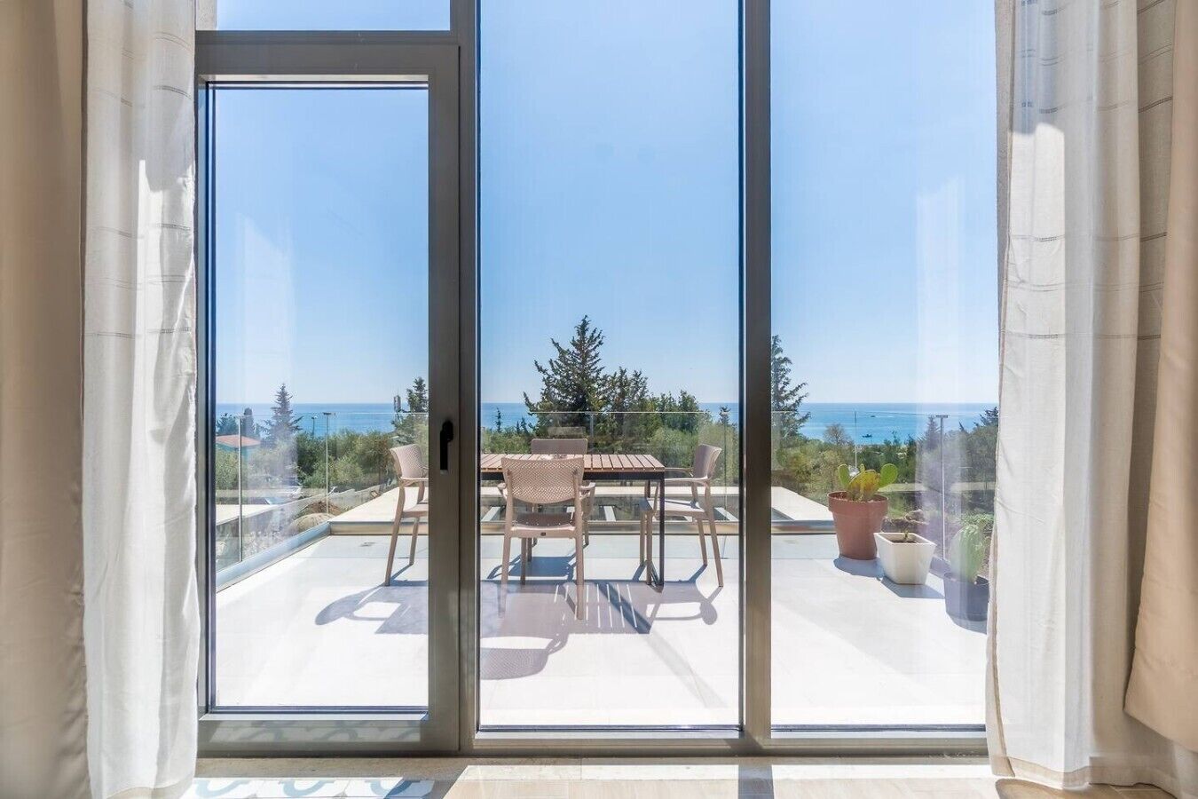 A view through a sliding glass door to an outdoor patio with a table and chairs, overlooking a coastal landscape.