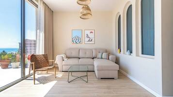 Uma sala de estar moderna com um sofá, uma mesa de centro de vidro e uma poltrona de vime. Há duas obras de arte emolduradas na parede, e o quarto tem grandes janelas com vista para o mar.