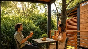Zwei Personen sitzen an einem Holztisch auf einer Terrasse, im Hintergrund ein Blumentopf und ein Schild mit der Aufschrift 'SHI'.