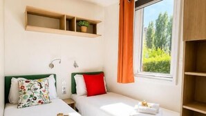 A hotel room with two beds, a window with curtains, a shelf above the beds, and a bedside table with a lamp.