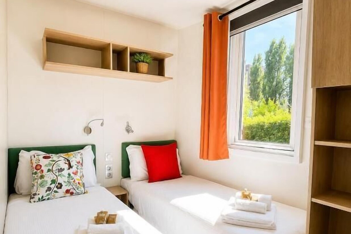 Ein Hotelzimmer mit zwei Betten, einem Fenster mit Vorhängen, einem Regal über den Betten und einem Nachttisch mit Lampe.