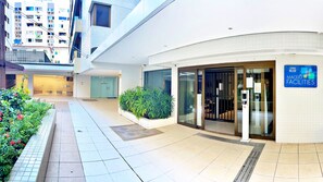 A modern building with a glass entrance, a sign for 'Maceió Facilities,' and a tiled pathway with plants. - MF102 | 3-bedroom, 3-bathroom apartment near Ponta Verde beach (Maceió)