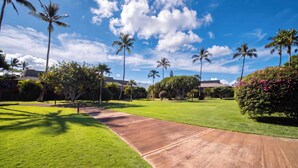 A well-maintained grassy area with a wooden pathway, surrounded by palm trees and lush greenery. - Koa Resort 3E | Two Bedroom Condo in Kihei (Kihei)