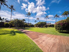 A well-maintained grassy area with a wooden pathway, surrounded by palm trees and lush greenery. - Koa Resort 3E | Two Bedroom Condo in Kihei (Kihei)