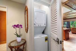 A laundry room with a washing machine, a small table with a potted orchid, and a door leading to a living area with a view of greenery. - Koa Resort 3E | Two Bedroom Condo in Kihei (Kihei)