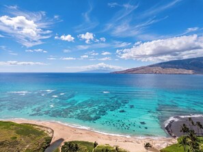 Un paisaje costero con aguas turquesas cristalinas, una playa arenosa y una cadena montañosa distante.