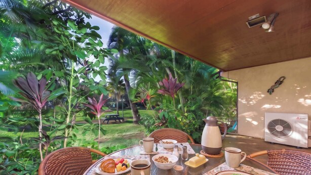 An outdoor dining area with wicker chairs, a table set with breakfast, and lush tropical vegetation in the background. - Koa Resort 3E | Two Bedroom Condo in Kihei (Kihei)