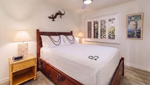 A bedroom with a bed, bedside lamps, a ceiling fan, a wall-mounted lizard decoration, a window with shutters, and a framed painting. - Koa Resort 3E | Two Bedroom Condo in Kihei (Kihei)