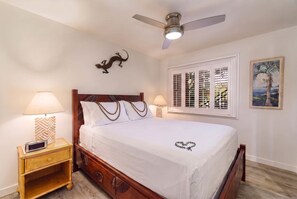 A bedroom with a bed, bedside lamps, a ceiling fan, a wall-mounted lizard decoration, a window with shutters, and a framed painting. - Koa Resort 3E | Two Bedroom Condo in Kihei (Kihei)