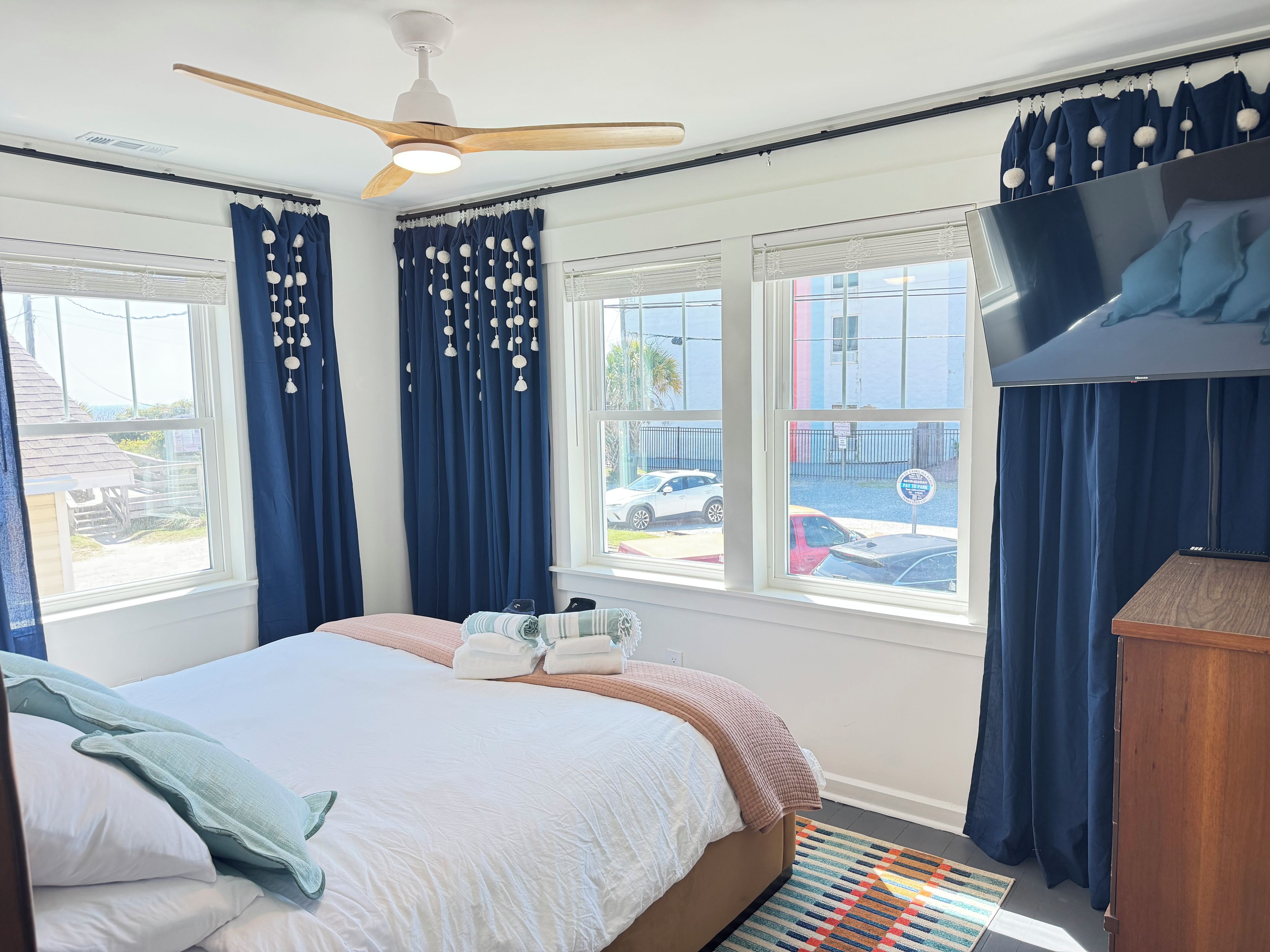 A hotel room with a bed, a ceiling fan, a TV, and large windows with curtains.