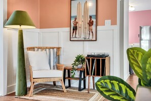 Ein Zimmer mit einem Korbstuhl, einer Stehlampe, einem gerahmten Bild von Surfern, einem kleinen Tisch mit einer Pflanze und einem Plattenspieler.
