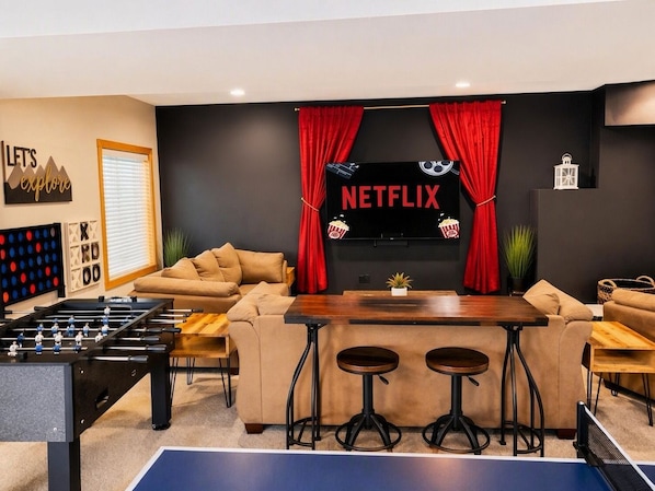 Una sala de estar con mesa de futbolín, mesa de ping pong, sofá y una pantalla de televisión que muestra el logotipo de Netflix.