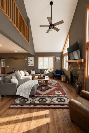 A spacious living room with a high ceiling, a fireplace, a large window, and a ceiling fan.