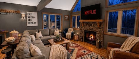Ein gemütliches Wohnzimmer mit Kamin, einem Netflix-Schild und einem großen Fenster mit Blick auf Bäume.