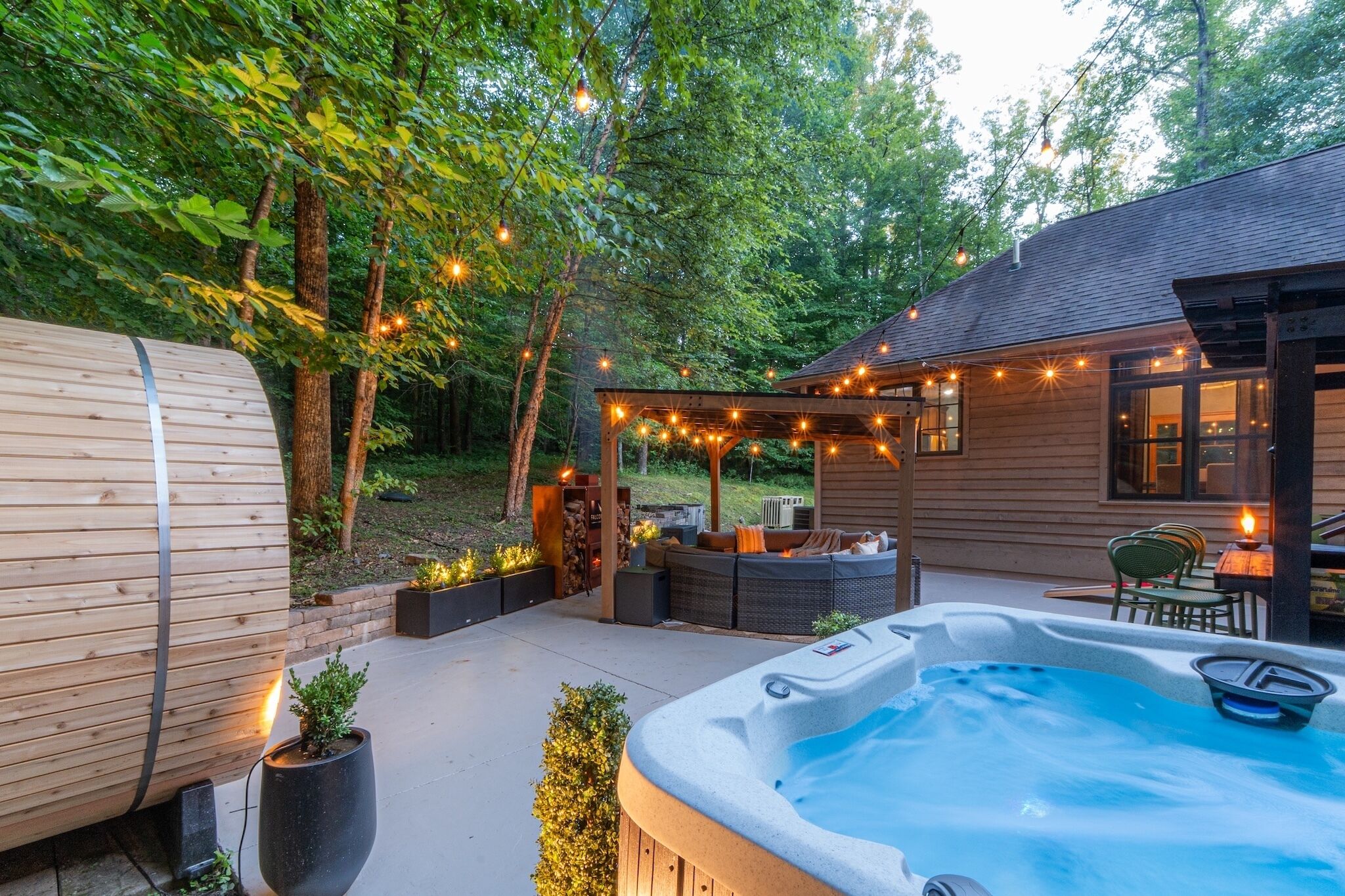 Un patio trasero con jacuzzi, terraza de madera, mobiliario exterior e iluminación con luces colgantes.