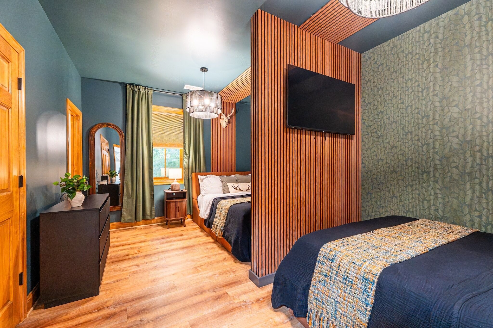 Een moderne hotelkamer met een houten vloer, een bed met een bed met een patroon, een bureau met een plant, een spiegel en een televisie aan de muur.