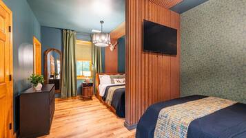 Une chambre d’hôtel moderne avec un plancher en bois, un lit recouvert d’une couverture à motifs, un bureau agrémenté d’une plante, un miroir et un téléviseur fixé au mur.