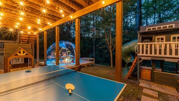 Une table de ping-pong extérieure, avec un filet, entourée de structures en bois et agrémentée d’un toboggan.