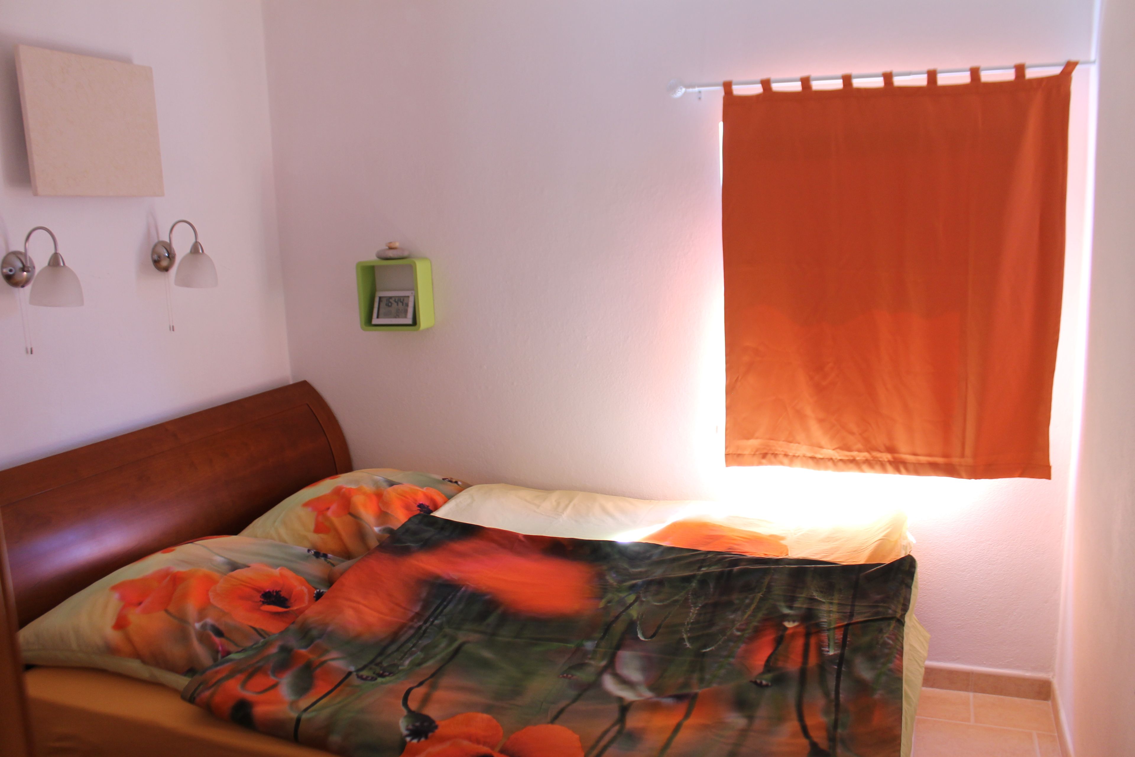 Een slaapkamer met een bed, nachtkastje, wandklok en raam met gordijn.