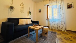 En stue med en sort sofa, et lille træbord, en gulvtæppe med mønster og et vindue med gardin med et træmotiv.