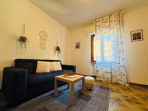 Ein Wohnzimmer mit einer schwarzen Couch, einem kleinen Holztisch, einem gemusterten Teppich und einem Vorhang mit Baummotiv.