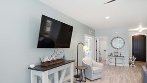 A modern living room with a flat-screen TV mounted on the wall, a white armchair, a small side table, and a round mirror on the wall. - New 4BR Ground-Level Home | Pool | Near Beach (Saint George Island)