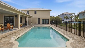 A swimming pool with a tiled deck, a covered patio area with chairs, and a two-story building in the background. - New 4BR Ground-Level Home | Pool | Near Beach (Saint George Island)
