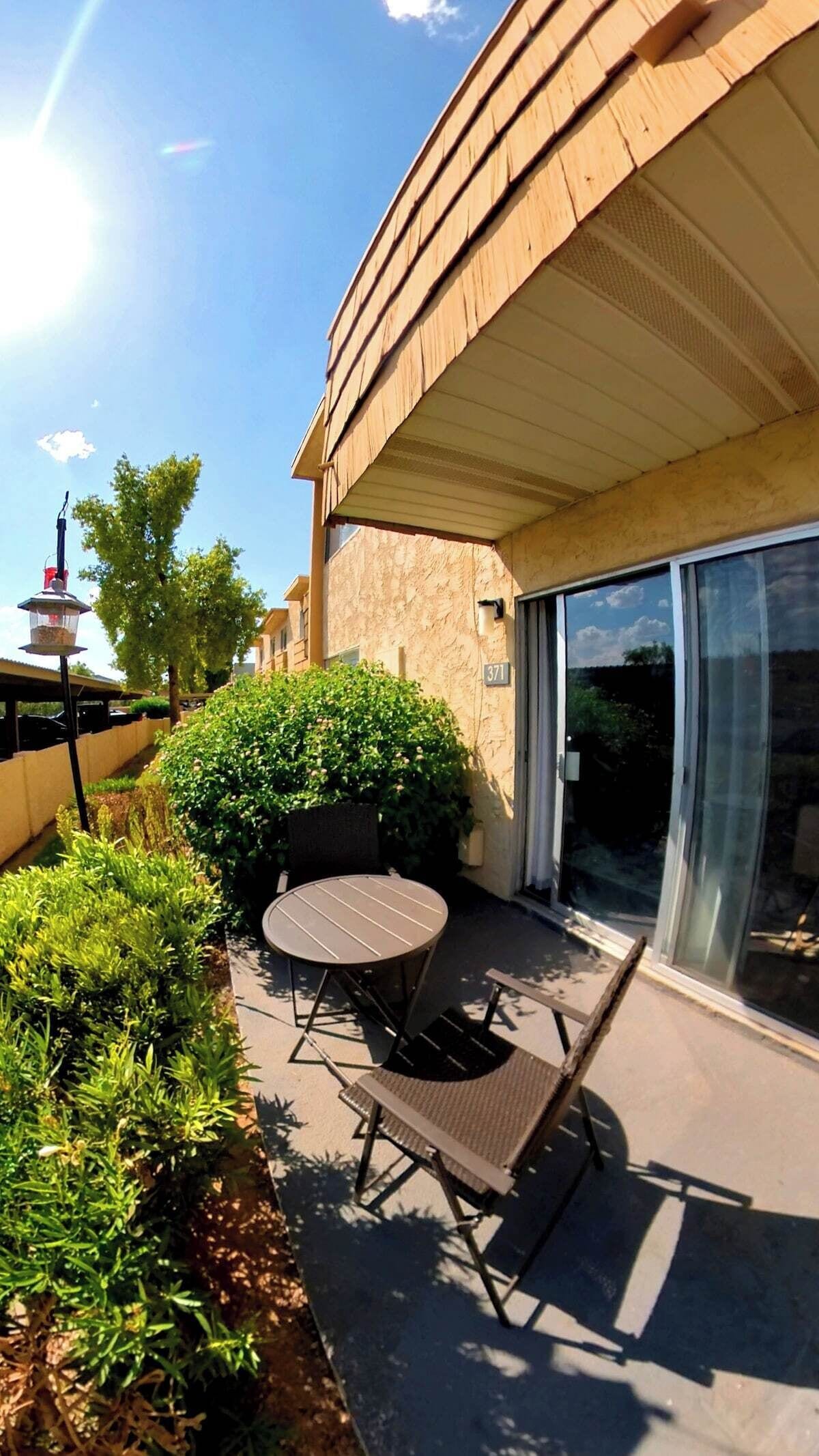 A covered outdoor seating area with a table and chairs, a potted plant, and a building with a glass door.