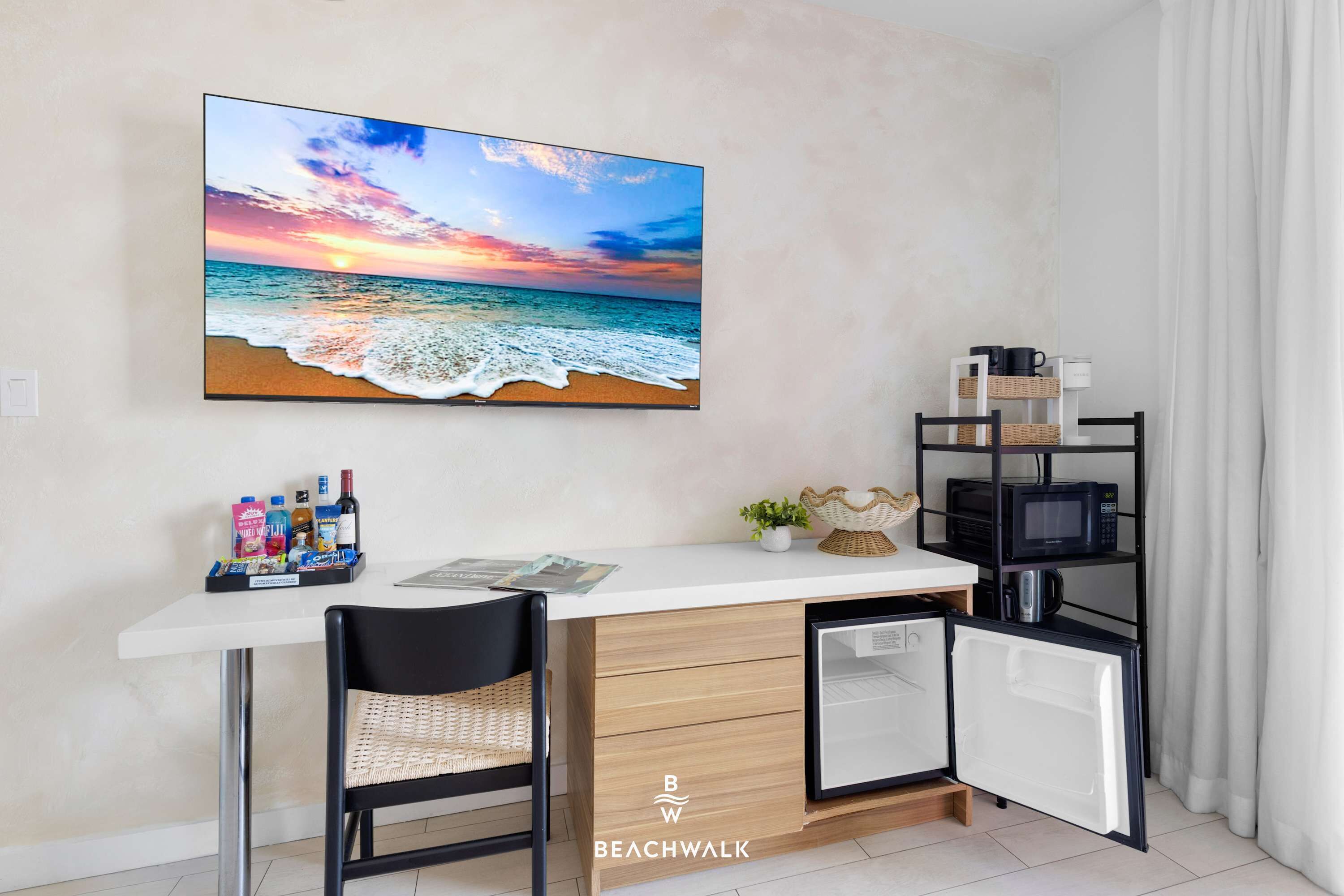 Il s’agit d’une pièce de taille modeste, comprenant un bureau blanc, une chaise, un petit réfrigérateur, un four à micro-ondes et une étagère où sont rangés des ustensiles de cuisine. Un grand tableau représentant une scène de plage orne le mur.
