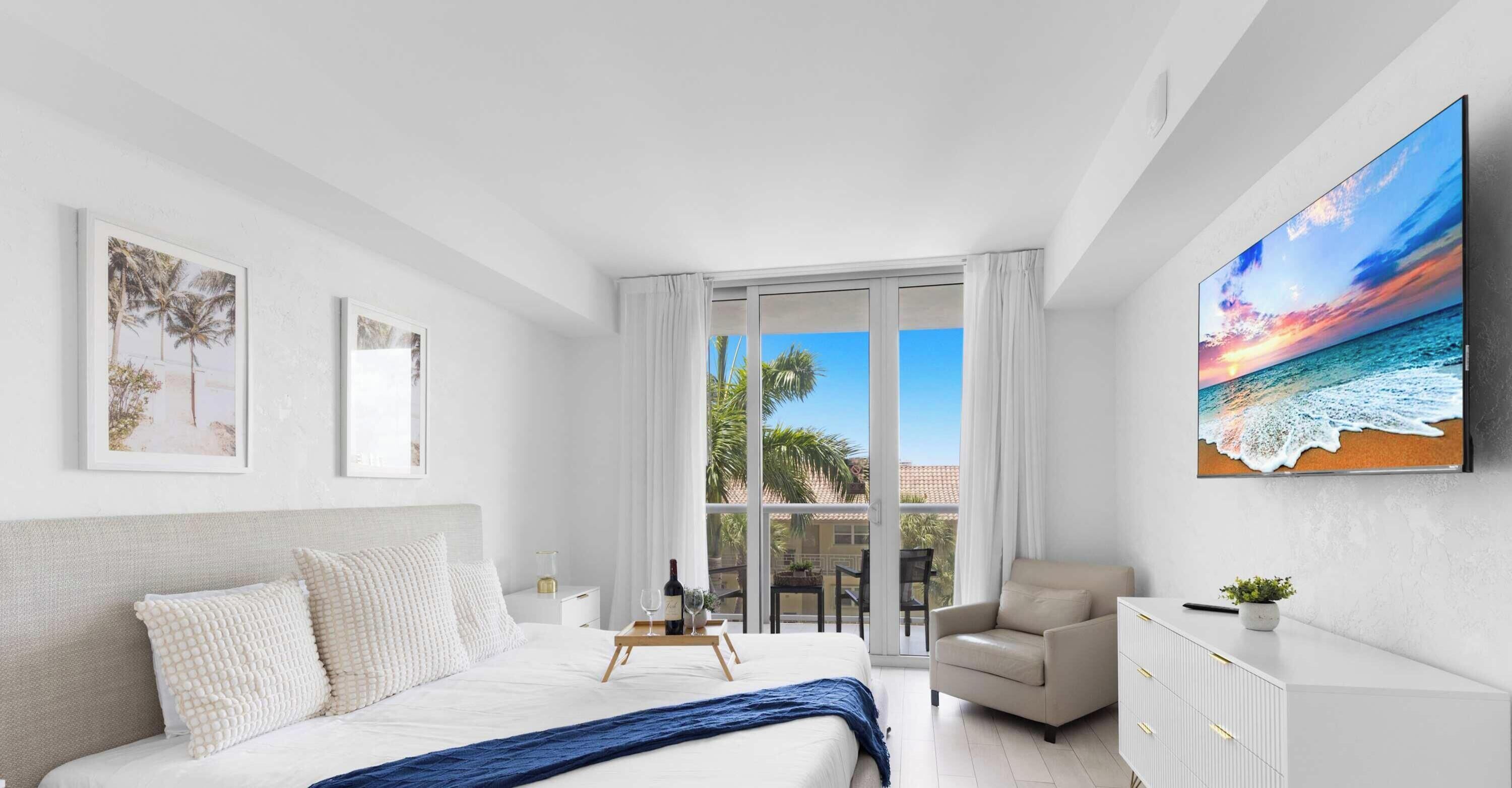A modern hotel room with a bed, a seating area, a dining table with wine, and a large window with a view of palm trees and the sea.