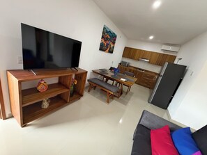 A modern living room with a flat-screen TV, wooden entertainment unit, dining table with chairs, and a sofa with cushions.