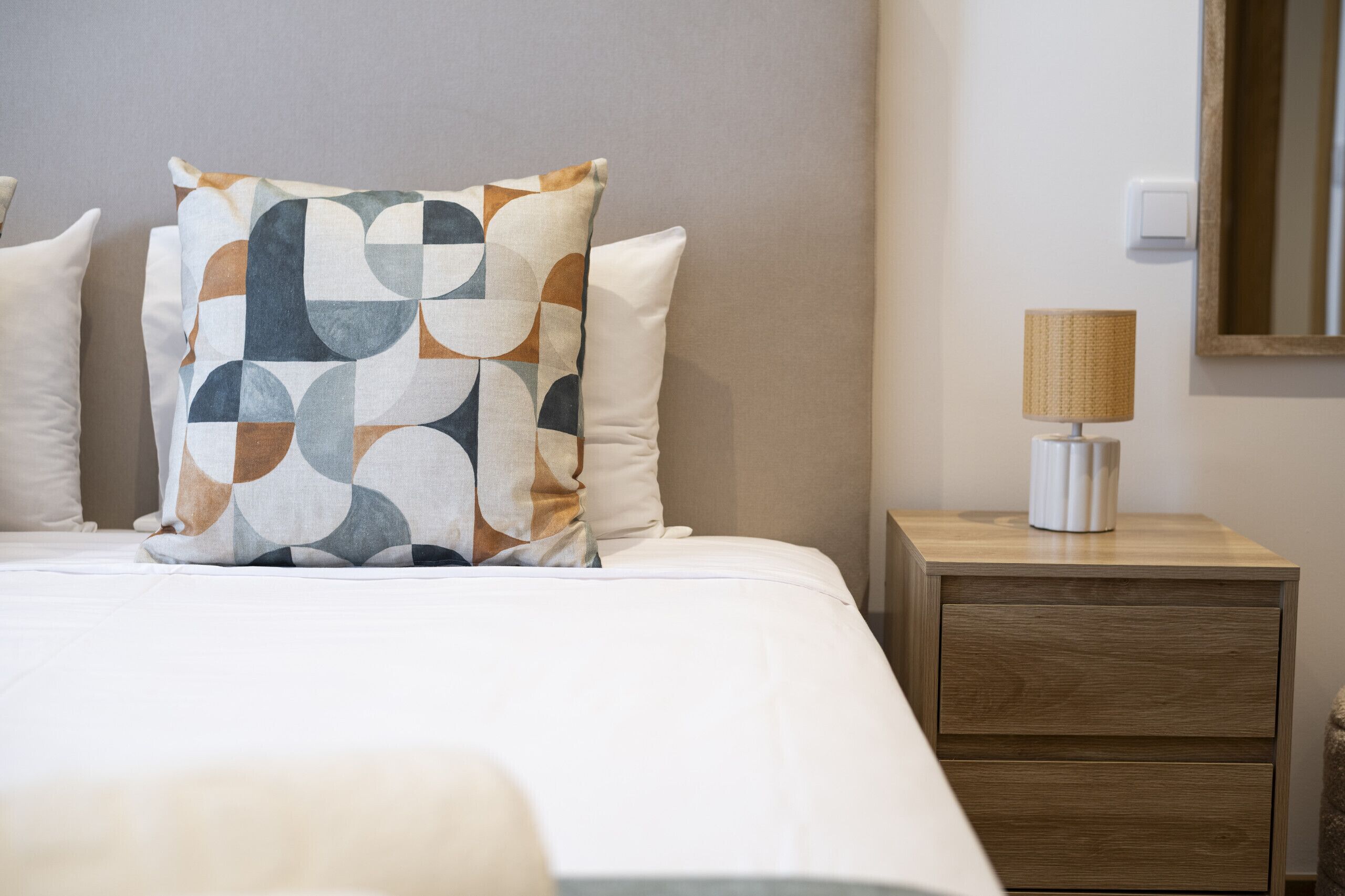 Un lit bien fait, agrémenté d’un coussin à motifs, d’une lampe de chevet et d’une table de nuit en bois dotée de tiroirs.