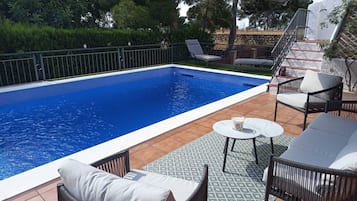 Une piscine rectangulaire dont le bord est revêtu de carreaux, entourée d’un patio aménagé avec du mobilier d’extérieur, notamment des chaises et une table.