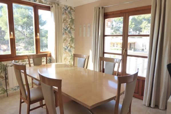 A dining area with a round table and chairs, large windows with curtains, and a view of the outdoors.