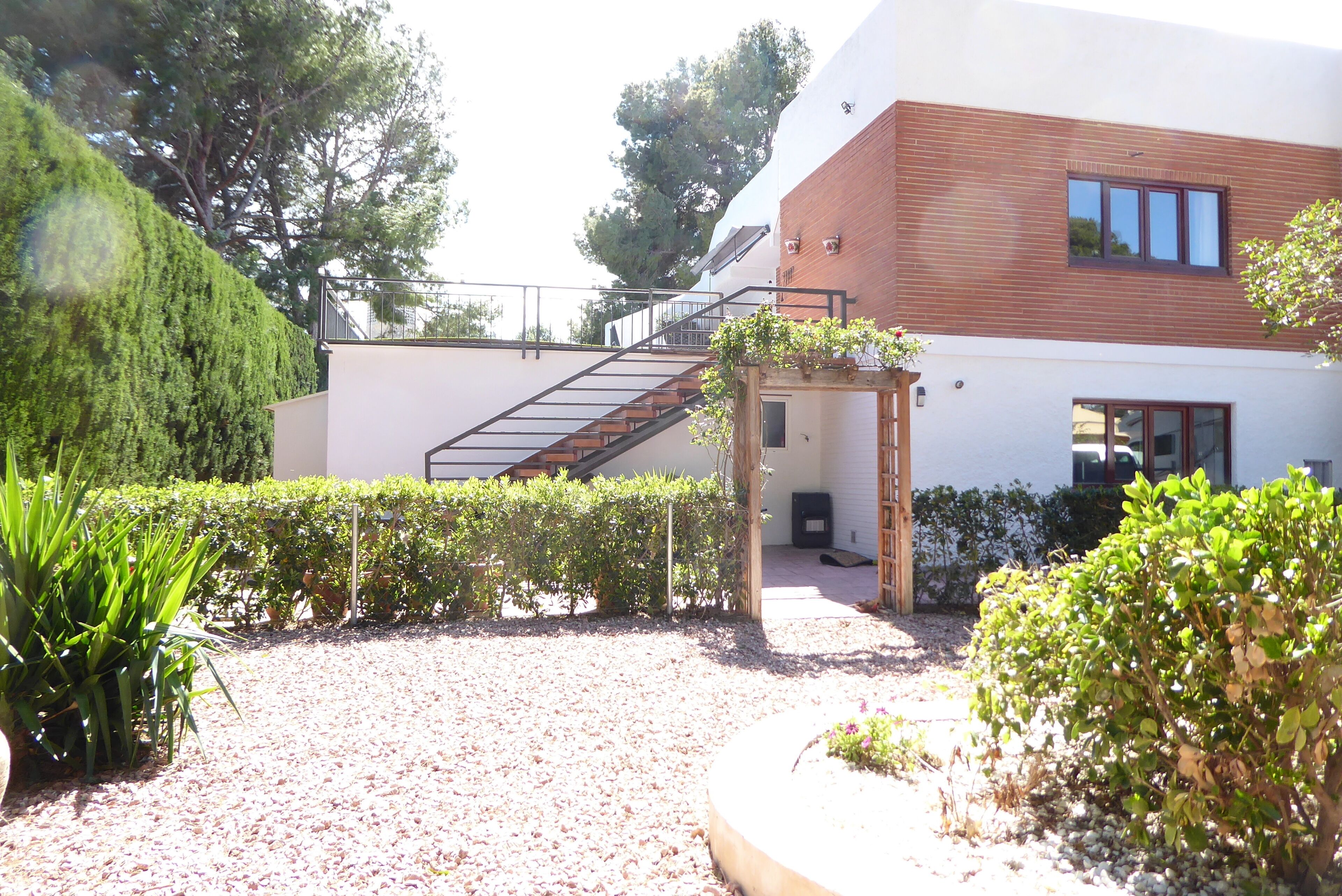 A modern building with a wooden staircase leading to a balcony, surrounded by well-maintained hedges and a gravel pathway.
