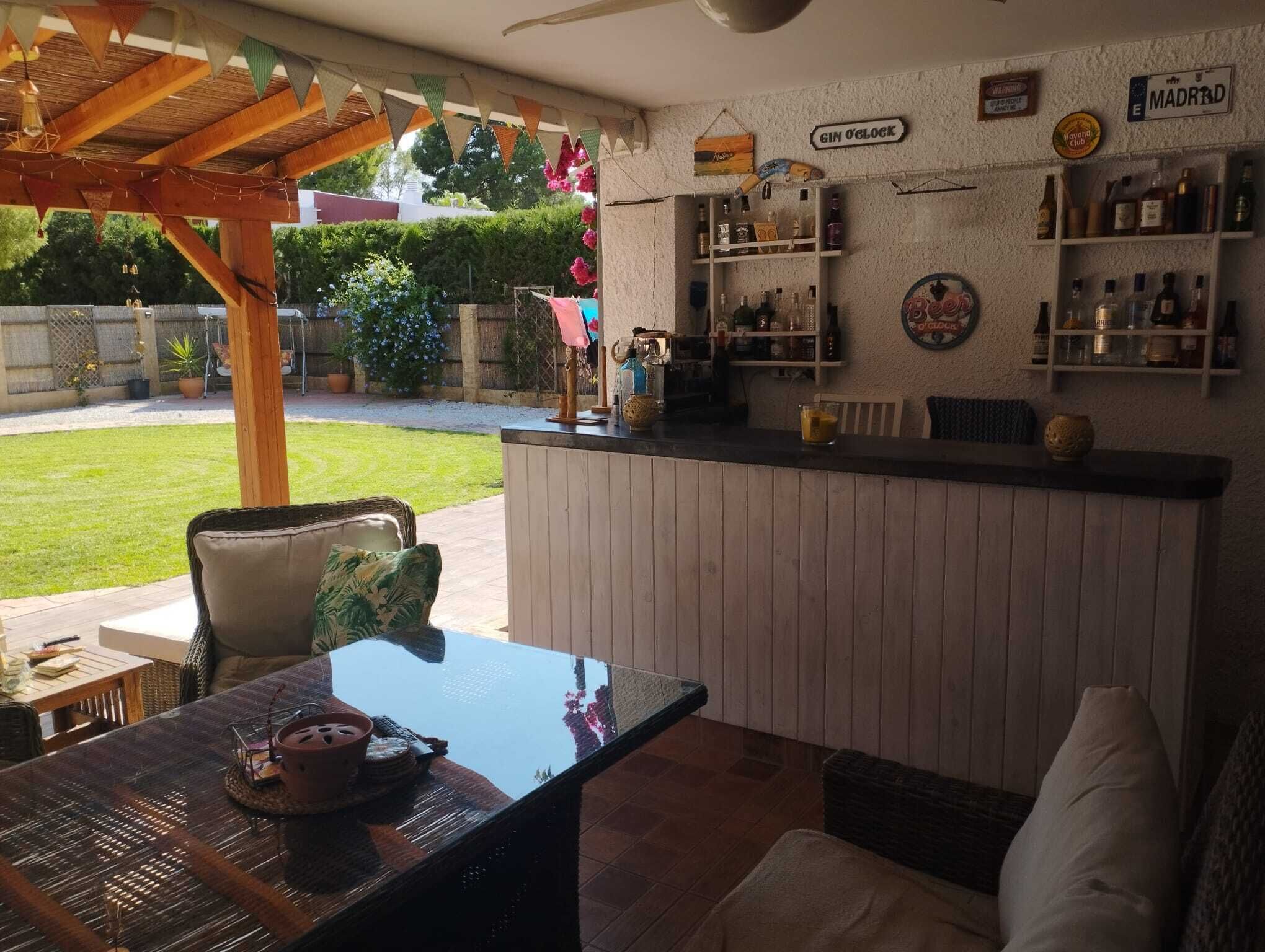 Een buitengebied met een bar, houten pergola, zitplaatsen en een bar met verschillende flessen en een bord waarop staat 'Gin O'Clock'.