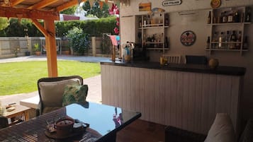 Een buitengebied met een bar, houten pergola, zitplaatsen en een bar met verschillende flessen en een bord waarop staat 'Gin O'Clock'.