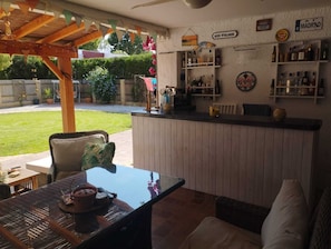 An outdoor bar area with a wooden pergola, seating, and a bar counter with various bottles and a sign reading 'Gin O'Clock'.