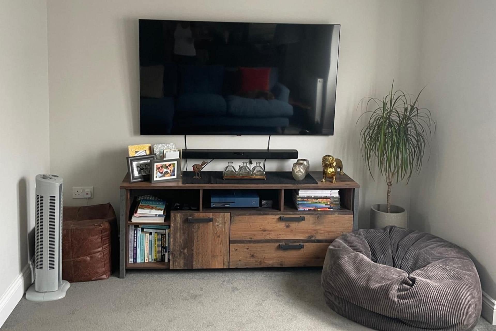 A living room with a flat-screen TV mounted on the wall, a wooden entertainment unit with books and decorative items, a standing fan, and a bean bag chair.