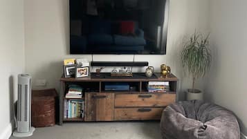 A living room with a flat-screen TV mounted on the wall, a wooden entertainment unit with books and decorative items, a standing fan, and a bean bag chair.