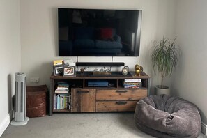 Ein Wohnzimmer mit einem an der Wand montierten Flachbildfernseher, einer Holzunterhaltungsecke mit Büchern und dekorativen Gegenständen, einem Standventilator und einem Sitzsack.