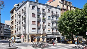 Um edifício com vários andares e varandas, uma rua em frente com bicicletas e pedestres, e áreas de estar ao ar livre.