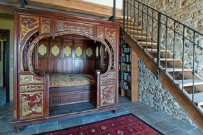 Ein traditionelles hölzernes Bett mit aufwendigen Schnitzereien, eine Steintreppe und ein Bücherregal voller Bücher.