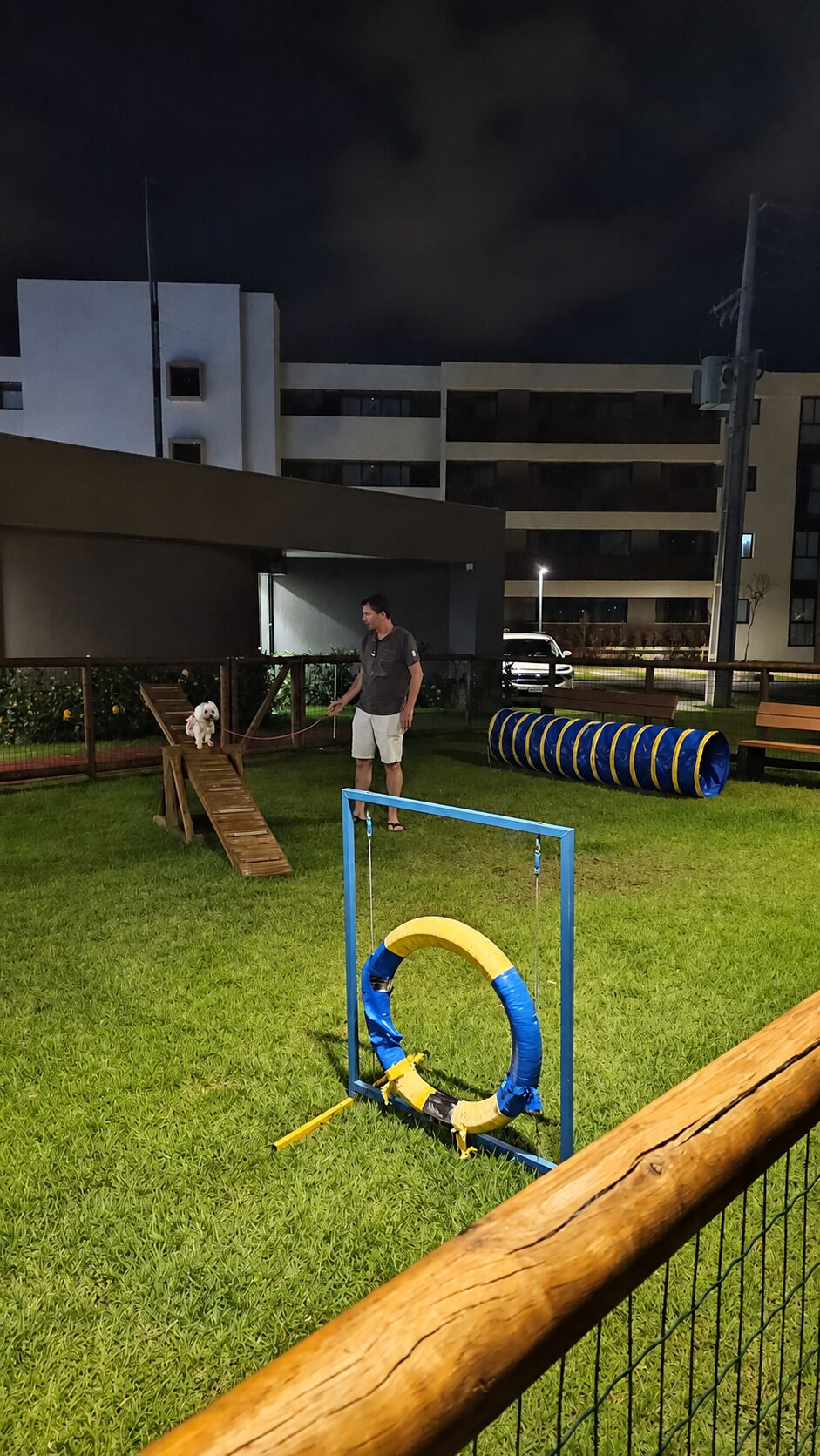Uma área ao ar livre com grama, cerca de metal, um banco de madeira e equipamento de exercícios azul e amarelo.