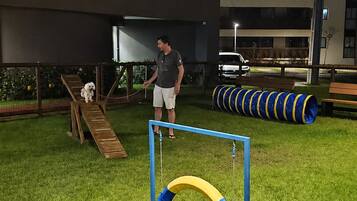 Uma área ao ar livre com grama, cerca de metal, um banco de madeira e equipamento de exercícios azul e amarelo.
