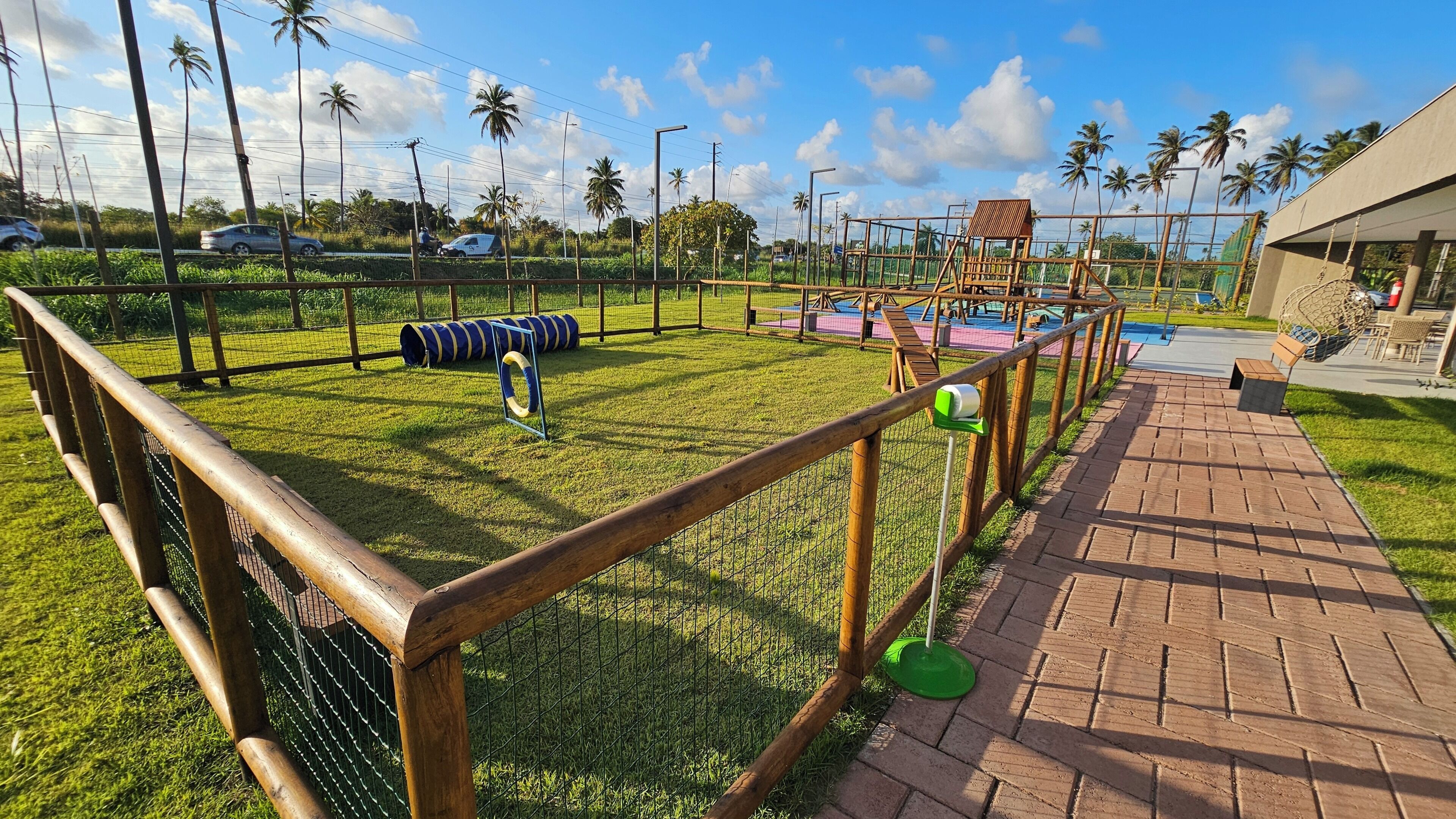 Un espacio exterior cercado con un parque infantil, un camino de ladrillo y palmeras al fondo.