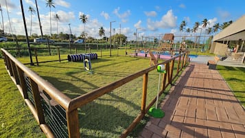 Un espacio exterior cercado con un parque infantil, un camino de ladrillo y palmeras al fondo.