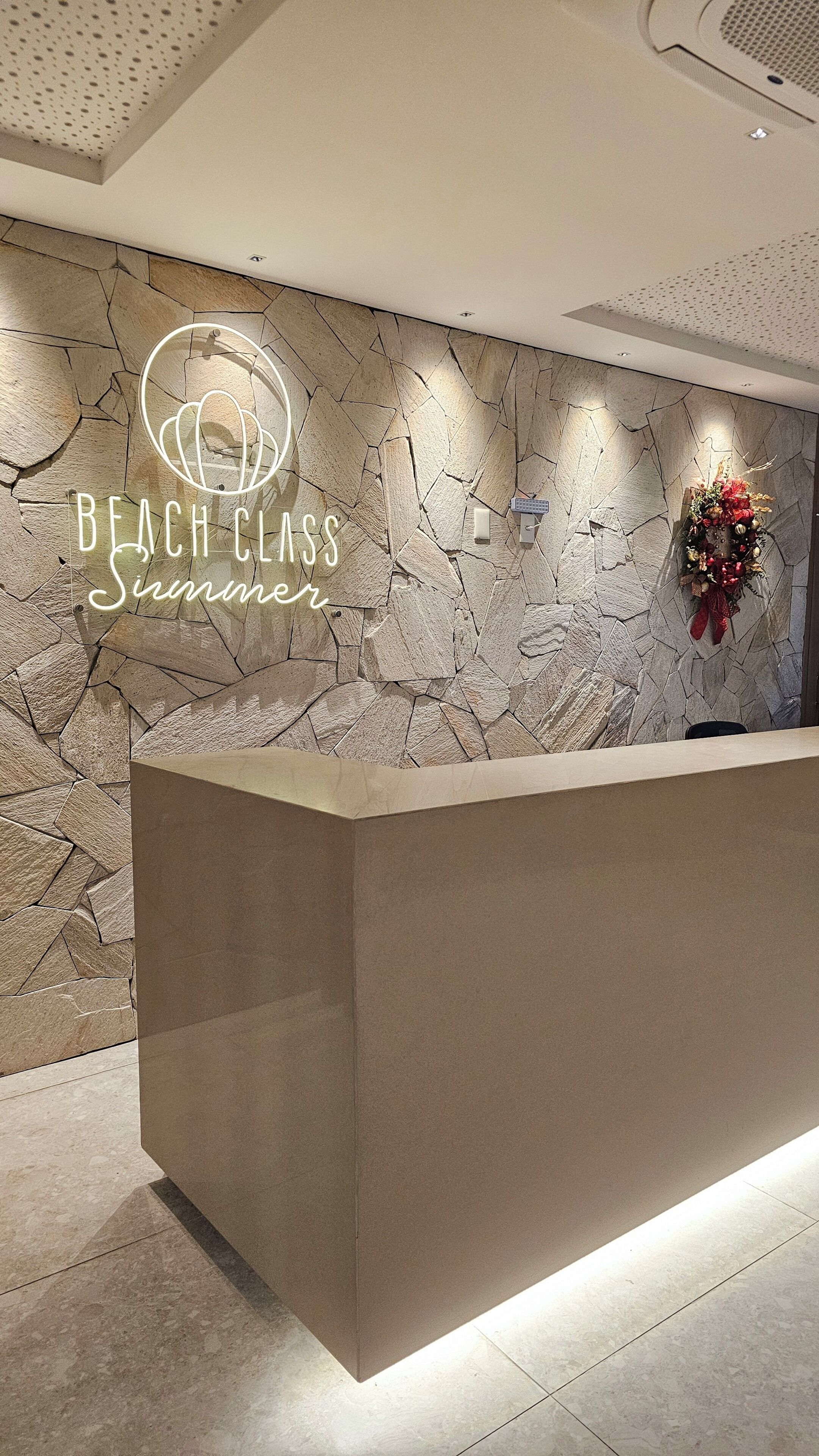 Un mostrador de recepción con un fondo de pared de piedra y un letrero de neón que dice 'BEACH CLASS Summer'.