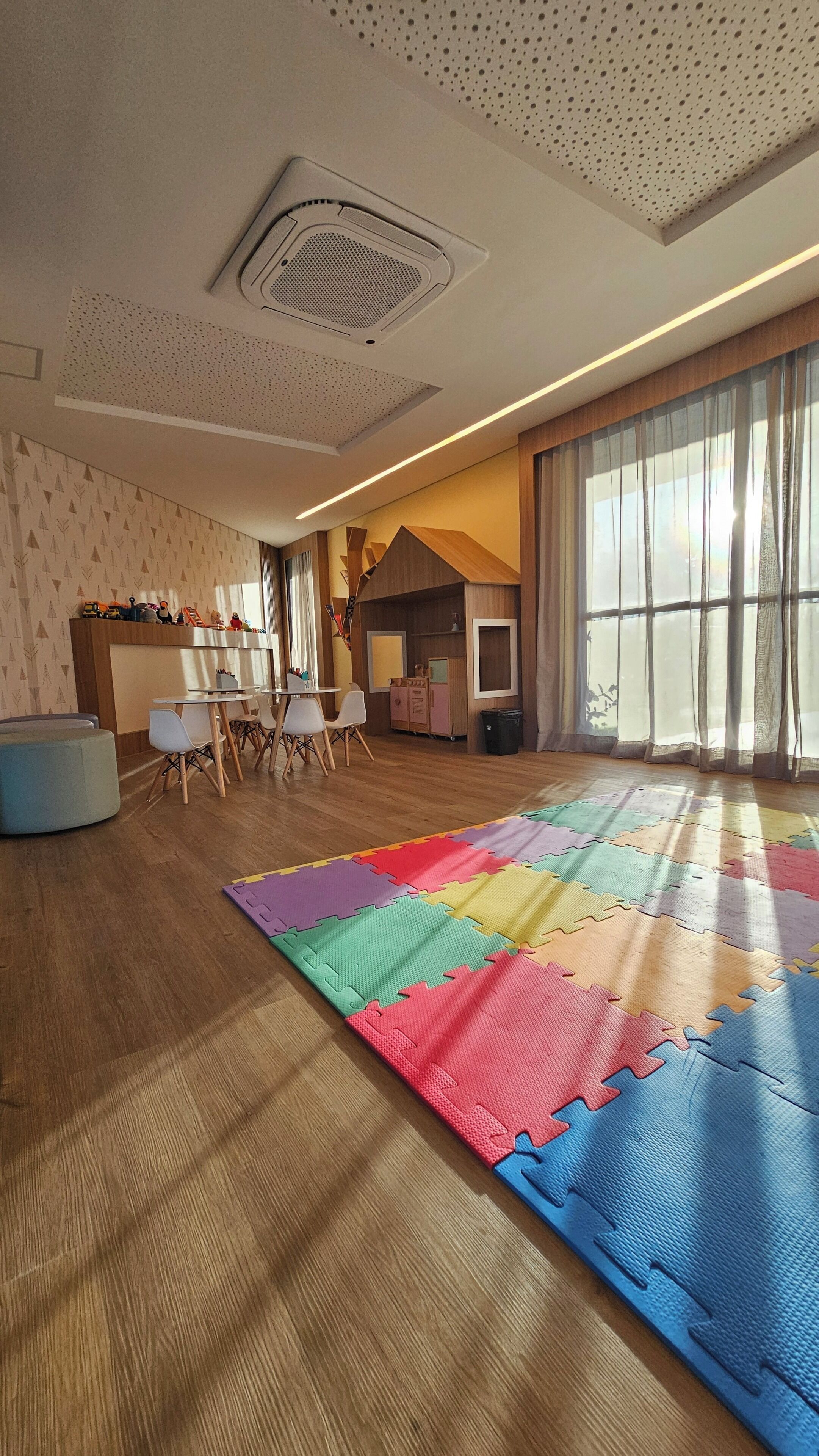 Um quarto espaçoso com uma grande janela, um respiradouro no teto, uma área de recreação com tapete de espuma colorido e uma cozinha de brinquedo infantil.